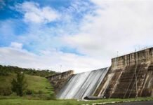 Barragem do Descoberto em sua capacidade máxima, cercada por vegetação exuberante e paisagens urbanas ao fundo, representando seu papel no abastecimento de cidades como Ceilândia e Taguatinga. O céu apresenta nuvens de chuva, simbolizando as recentes precipitações que levaram ao aumento do nível da água.