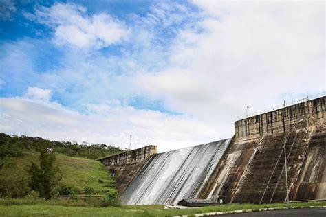 OIP (1) Barragem do Descoberto em sua capacidade máxima, cercada por vegetação exuberante e paisagens urbanas ao fundo, representando seu papel no abastecimento de cidades como Ceilândia e Taguatinga. O céu apresenta nuvens de chuva, simbolizando as recentes precipitações que levaram ao aumento do nível da água.