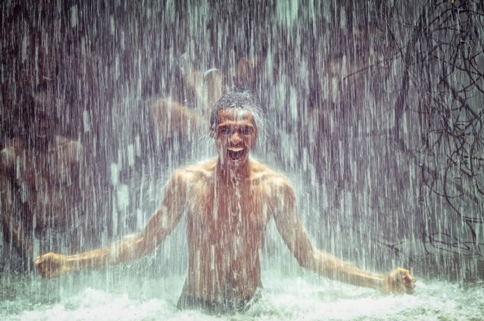 Homem tomando banho de cachoeira, feliz e imerso na natureza, com água caindo ao seu redor, representando a prática de Shinrin-yoku ou banho de floresta.