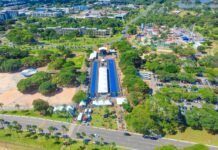 Vista aérea de parque urbano em Brasília durante evento ao ar livre, com tendas, estrutura coberta azul, parque de diversões e áreas verdes ao redor.