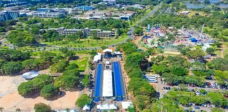 Vista aérea de parque urbano em Brasília durante evento ao ar livre, com tendas, estrutura coberta azul, parque de diversões e áreas verdes ao redor.