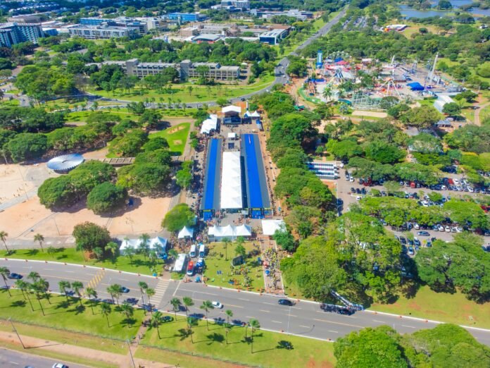 pexels-banco-de-imagens-sel-df-643358816-30404864 Vista aérea de parque urbano em Brasília durante evento ao ar livre, com tendas, estrutura coberta azul, parque de diversões e áreas verdes ao redor.