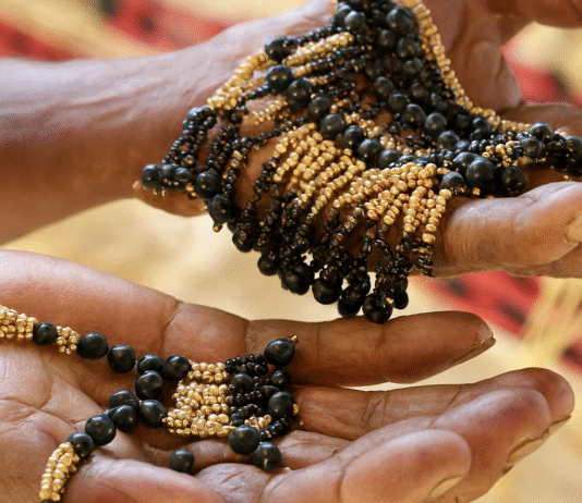 Mãos envelhecidas, de pele castanho-escura e rugosa, seguram colares artesanais feitos de pequenas sementes cilíndricas bege-douradas e contas pretas brilhantes, com fundo desfocado em tons terrosos e tecido vermelho e preto.