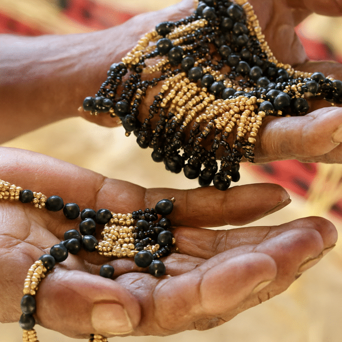 Mãos envelhecidas, de pele castanho-escura e rugosa, seguram colares artesanais feitos de pequenas sementes cilíndricas bege-douradas e contas pretas brilhantes, com fundo desfocado em tons terrosos e tecido vermelho e preto.